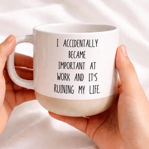 Pode incluir: Caneca de café de cerâmica branca com base bege e alça curva. A caneca apresenta o texto preto: "I ACCIDENTALLY BECAME IMPORTANT AT WORK AND IT'S RUINING MY LIFE."