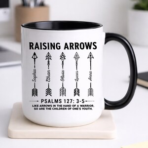 May include: A white ceramic mug with a black interior and handle. The front features the black text RAISING ARROWS above five vertical arrow illustrations, each labeled with the names Sophia, Ethan, Olivia, Lucas, and Anna. Below are the words PSALMS 127: 3-5 and the quote LIKE ARROWS IN THE HAND OF A WARRIOR, SO ARE THE CHILDREN OF ONE'S YOUTH.