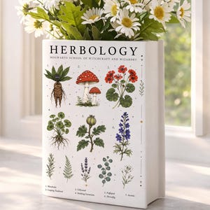 May include: A white rectangular ceramic vase featuring a botanical Herbology print from Hogwarts School of Witchcraft and Wizardry. The design displays numbered illustrations of various plants, including a mandrake and mushrooms, filled with white daisies.