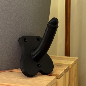 Könnte beinhalten: Eine schwarze, phallusförmige Wandskulptur. Die Skulptur ist auf einer rechteckigen Basis mit drei Schraubenlöchern befestigt. Die Skulptur besteht aus einem mattschwarzen Material und wird vor einem neutralen Hintergrund gezeigt.