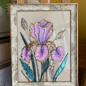Op de afbeelding: Ingelijst bloemkunstwerk met een gedetailleerde iris in paarse en gouden tinten. Het schilderij heeft een textuur, met turquoise en groene bladeren en stelen. De lijst is verweerd wit met gouden rand, wat een vintage esthetiek creëert.