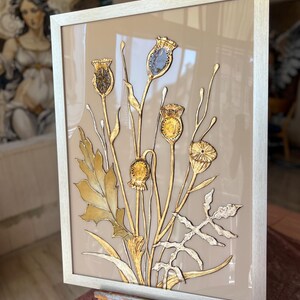 Op de afbeelding: Een ingelijst kunstwerk met een botanisch ontwerp met gouden en zilveren accenten. Het kunstwerk toont gestileerde bloemen en bladeren tegen een neutrale achtergrond, omlijst door een zilveren lijst. Het stuk wordt op een houten ezel getoond.