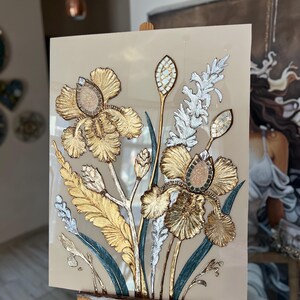 Op de afbeelding: Een bloemkunstwerk met gouden, zilveren en teal accenten. Het stuk toont gedetailleerde irissen en bladeren, op een beige achtergrond. Het kunstwerk wordt gepresenteerd op een houten ezel, wat de artistieke presentatie benadrukt.