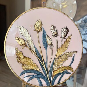Op de afbeelding: Ronde wandkunst met een bloemmotief en gouden en zilveren accenten. Het kunstwerk heeft een lichtroze achtergrond en een gouden lijst. De bloemen en bladeren zijn gedetailleerd met metallic goud en zilver, wat een driedimensionaal effect creëert.