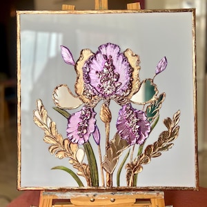 Op de afbeelding: Een ingelijst kunstwerk met een gedetailleerde irisbloem. De iris heeft paarse bloemblaadjes met gouden accenten, tegen een lichtgrijze achtergrond. De lijst is goudkleurig. Het kunstwerk wordt tentoongesteld op een houten ezel.