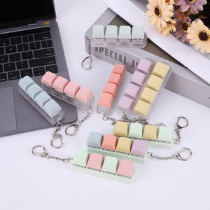 May include: A selection of pastel-coloured keyboard keycap keychains, featuring pink, blue, yellow, and green keycaps. Each keychain holds five keycaps and is attached to a silver keyring. A laptop is visible in the background.