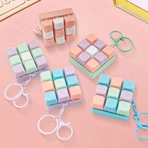 Puede incluir: Una colección de llaveros con teclas de teclado de colores pastel en tonos rosa, azul, verde y morado. Cada llavero presenta una cuadrícula de 3x3 teclas y un llavero de metal. Los llaveros están dispuestos sobre una superficie rosa.