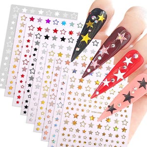 May include: Nail art stickers featuring star designs in various colors, including red, black, gold, and silver. The image also shows several long, pointed nails decorated with star-shaped embellishments and rhinestones.