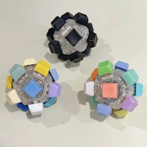 Könnte beinhalten: Drei kugelförmige, geometrische Objekte mit farbigen Tastenkappen. Eines ist schwarz, die anderen sind mehrfarbig mit Pastelltönen. Jedes hat eine zentrale quadratische Tastenkappe und einen klaren, facettierten Körper.