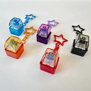 以下が含まれることがあります： 青、オレンジ、紫、赤、黄、黒など、さまざまな色のカラフルなキーキャップチャームのコレクション。各チャームは、星型のキーチェーンアタッチメントを備えた透明な正方形のキーキャップデザインが特徴です。キーキャップは十字型のデザインです。
