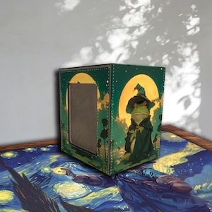 May include: A green and yellow card storage box featuring a cartoon frog dressed as a wizard. The frog is wearing a green hat and cloak, set against a backdrop of a full moon and trees. The box has a rectangular opening on one side.
