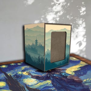Peut inclure: Une boîte de rangement carrée pour cartes avec un paysage de montagnes, d'arbres et une pagode dans des tons de bleu et de vert. La boîte a une découpe rectangulaire sur le devant et est posée sur un tapis coloré.