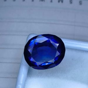 May include: A faceted, oval-shaped, deep blue gemstone. The gem has multiple facets that reflect light, creating a brilliant sparkle. The stone is set against a light background, highlighting its color and clarity. The gem is likely a sapphire or similar precious stone.