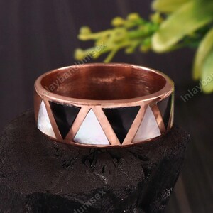 Può includere: Un anello in rame caratterizzato da un motivo geometrico ripetitivo di intarsi triangolari alternati in nero e bianco iridescente. La fascia presenta una finitura metallica liscia e lucida, con una larghezza costante e un design pulito.