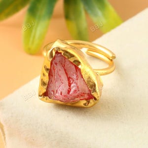 May include: A gold-toned adjustable ring featuring a raw, triangular-shaped pink gemstone with red veining. The gemstone is set in a textured gold bezel. The ring is displayed on a cream-colored surface, with blurred green and yellow foliage in the background.