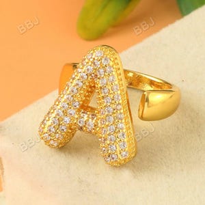 Könnte beinhalten: Ein goldfarbener, verstellbarer Ring mit dem Buchstaben "A". Der Buchstabe ist mit kleinen, klaren Edelsteinen besetzt. Der Ring hat eine polierte Oberfläche und ist vor einem neutralen Hintergrund platziert.