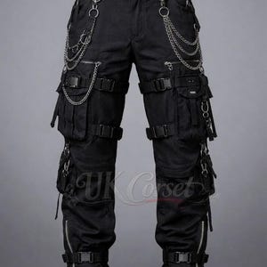 Puede incluir: Pantalones cargo negros con múltiples bolsillos, correas y detalles de cadenas plateadas. Los pantalones tienen cremalleras en los tobillos y un cinturón con hebilla. El logo de UK Corset es visible.