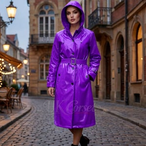 Puede incluir: Un impermeable morado brillante con capucha, cinturón y botones. El abrigo largo tiene un cinturón con hebilla en la cintura y está hecho de un material brillante. El texto "UK Corset" es visible.