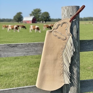 May include: A tan leather chaps with decorative stitching and fringe, hanging on a wooden post. The chaps have a brown leather strap. In the background, a wooden fence, green field, and grazing cows are visible.