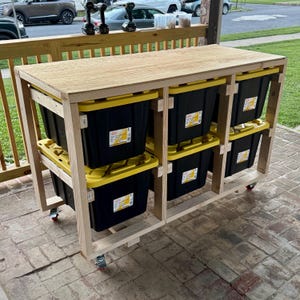 May include: A wooden storage cart with six black and yellow storage bins. The cart has a wooden top and is on wheels. The cart is made of light-coloured wood and is outdoors.