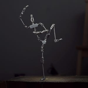 May include: A silver articulated figure in a dynamic pose, with multiple joints and a detailed mechanical design. The figure is positioned on a textured surface against a dark background, suggesting a studio setting. The figure's arms and legs are extended.