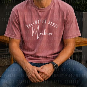 Puede incluir: Camiseta de color rosa polvoriento con el texto blanco "SALTWATER REBEL Mockups". El modelo lleva vaqueros azules y un reloj negro. La camiseta tiene cuello redondo y mangas cortas.