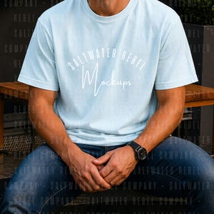 Puede incluir: Una camiseta azul claro con el texto "Saltwater Rebel Mockups" en escritura blanca. La camiseta se combina con vaqueros azules y un reloj negro. La persona está sentada en un banco de madera.