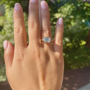 May include: A gold ring featuring a large, rectangular, pale gemstone. The ring is displayed on a hand with manicured nails. The background is blurred, with green foliage visible.