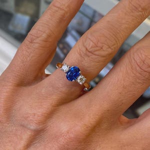 May include: A gold ring with a large oval blue gemstone in the center, flanked by two smaller, round, clear gemstones. The ring is set on a finger, showcasing the design and sparkle of the jewelry.