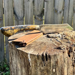 Puede incluir: Un cuchillo artesanal con mango de madera con vetas amarillas y negras. La hoja plateada descansa sobre una peque&ntilde;a funda de madera rectangular. El cuchillo se exhibe sobre un toc&oacute;n de &aacute;rbol desgastado.