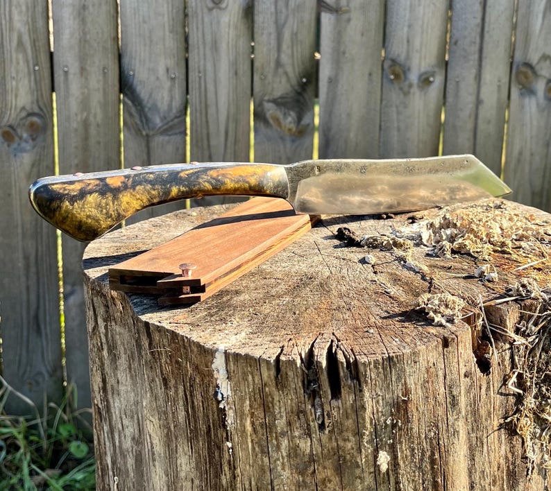 Puede incluir: Un cuchillo artesanal con mango de madera con vetas amarillas y negras. La hoja plateada descansa sobre una peque&ntilde;a funda de madera rectangular. El cuchillo se exhibe sobre un toc&oacute;n de &aacute;rbol desgastado.