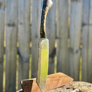 Puede incluir: Un cuchillo artesanal con mango de madera y hoja de acero inoxidable. El mango presenta un dise&ntilde;o oscuro y estampado, mientras que la hoja tiene una superficie ligeramente texturizada. El cuchillo est&aacute; en una funda de madera sobre un toc&oacute;n.