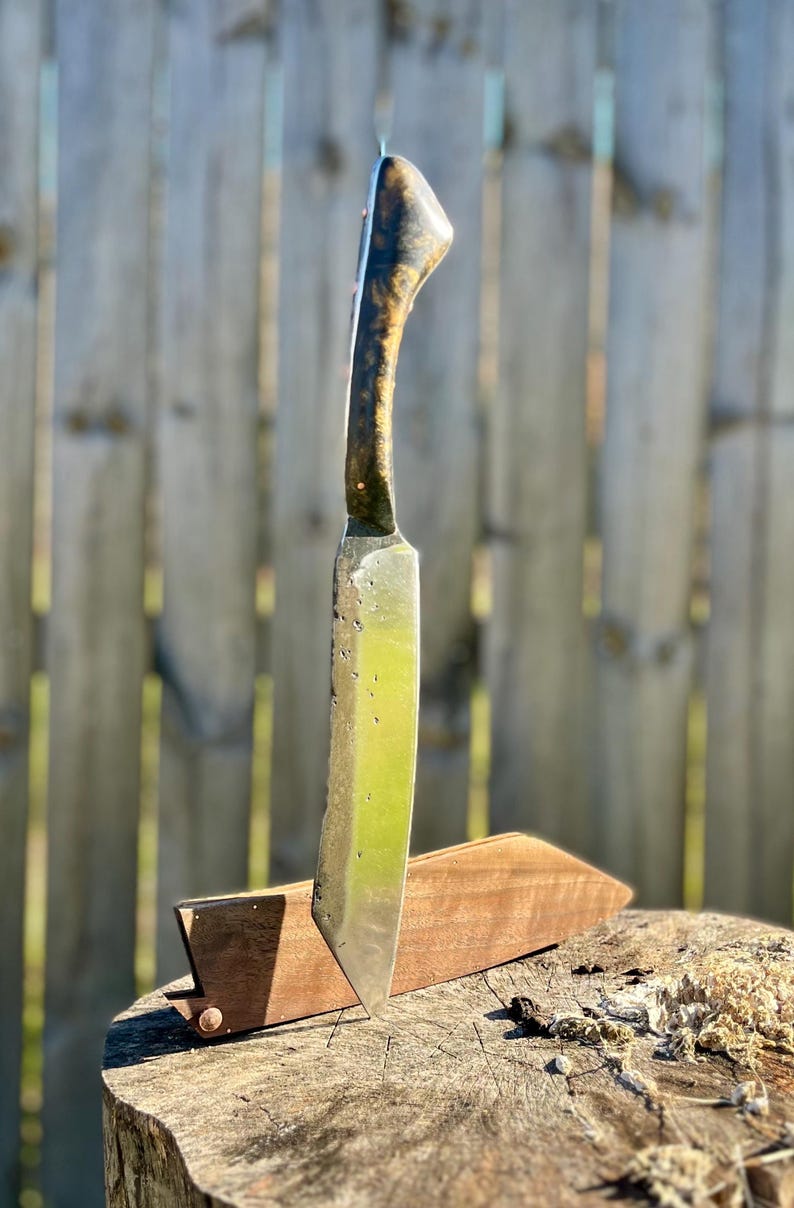 Puede incluir: Un cuchillo artesanal con mango de madera y hoja de acero inoxidable. El mango presenta un dise&ntilde;o oscuro y estampado, mientras que la hoja tiene una superficie ligeramente texturizada. El cuchillo est&aacute; en una funda de madera sobre un toc&oacute;n.