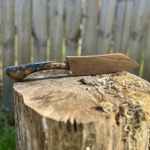 Puede incluir: Un cuchillo de chef artesanal con un mango de madera con un patr&oacute;n veteado &uacute;nico en tonos marr&oacute;n, negro y dorado. La hoja est&aacute; protegida por una funda de madera. El cuchillo descansa sobre un toc&oacute;n de &aacute;rbol.