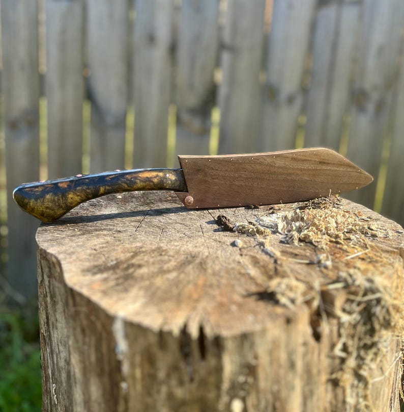 Puede incluir: Un cuchillo de chef artesanal con un mango de madera con un patr&oacute;n veteado &uacute;nico en tonos marr&oacute;n, negro y dorado. La hoja est&aacute; protegida por una funda de madera. El cuchillo descansa sobre un toc&oacute;n de &aacute;rbol.
