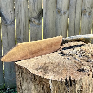 Puede incluir: Un cepillo de madera vintage con un mango de metal oscuro descansa sobre un toc&oacute;n de &aacute;rbol. El cuerpo de madera del cepillo es de un marr&oacute;n c&aacute;lido, y el mango tiene un aspecto deslustrado y envejecido. La herramienta est&aacute; dise&ntilde;ada para carpinter&iacute;a.