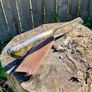 Puede incluir: Una navaja recta con una hoja de metal pulido y un mango oscuro con remaches de cobre descansa sobre una correa de cuero de madera. La navaja y la correa se colocan sobre un toc&oacute;n de &aacute;rbol desgastado, lo que sugiere una est&eacute;tica r&uacute;stica o vintage.