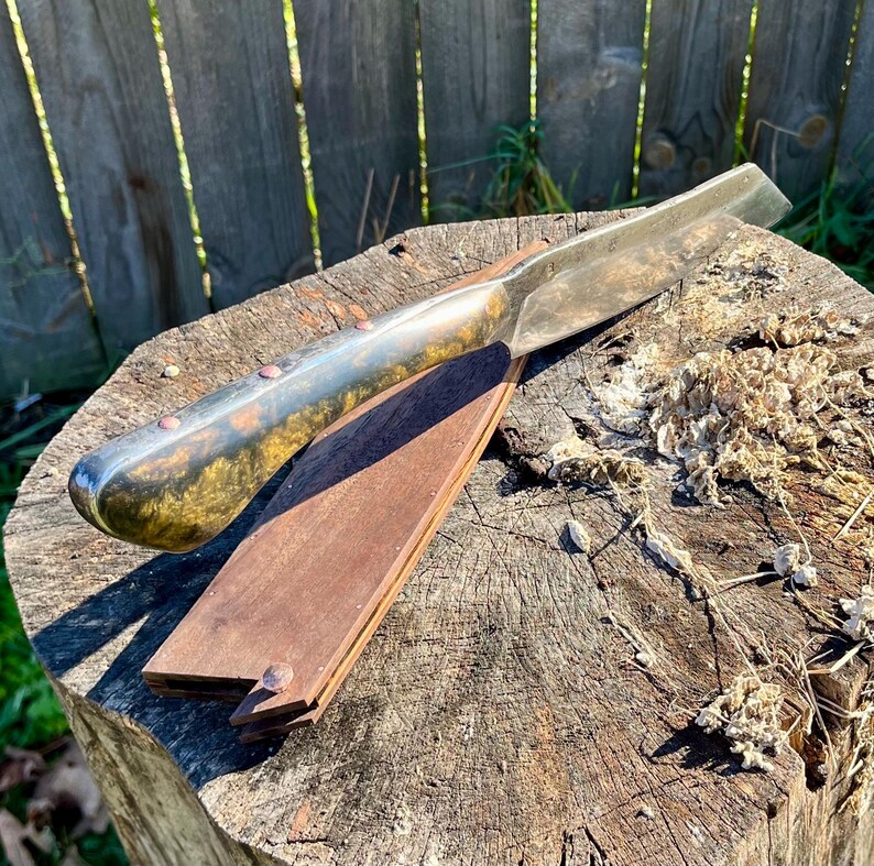 Puede incluir: Una navaja recta con una hoja de metal pulido y un mango oscuro con remaches de cobre descansa sobre una correa de cuero de madera. La navaja y la correa se colocan sobre un toc&oacute;n de &aacute;rbol desgastado, lo que sugiere una est&eacute;tica r&uacute;stica o vintage.