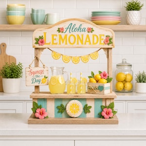 May include: A wooden lemonade stand decoration featuring the text Aloha LEMONADE and Squeeze the Day. The stand includes a glass pitcher of yellow lemonade, two glass mugs with yellow striped straws, a crate of lemons, and pink hibiscus flowers.