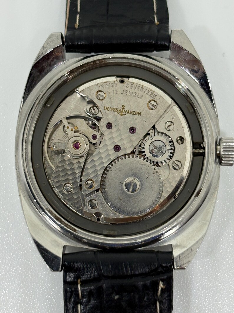 K&ouml;nnte beinhalten: Eine silberfarbene Armbanduhr mit abgenommenem Geh&auml;useboden, die das mechanische Uhrwerk freilegt. Das Werk ist mit Ulysse Nardin, Swiss und Seventeen 17 Jewels graviert. Sie hat ein schwarzes Lederarmband mit wei&szlig;en Ziern&auml;hten.