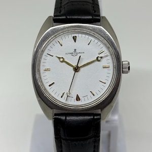 K&ouml;nnte beinhalten: Eine silberfarbene Armbanduhr mit wei&szlig;em Zifferblatt, goldfarbenen Zeigern und einem Anker-Logo &uuml;ber dem Schriftzug Ulysse Nardin. Die Uhr verf&uuml;gt &uuml;ber ein schwarzes Lederarmband mit strukturierter Oberfl&auml;che und eine silberne Krone.
