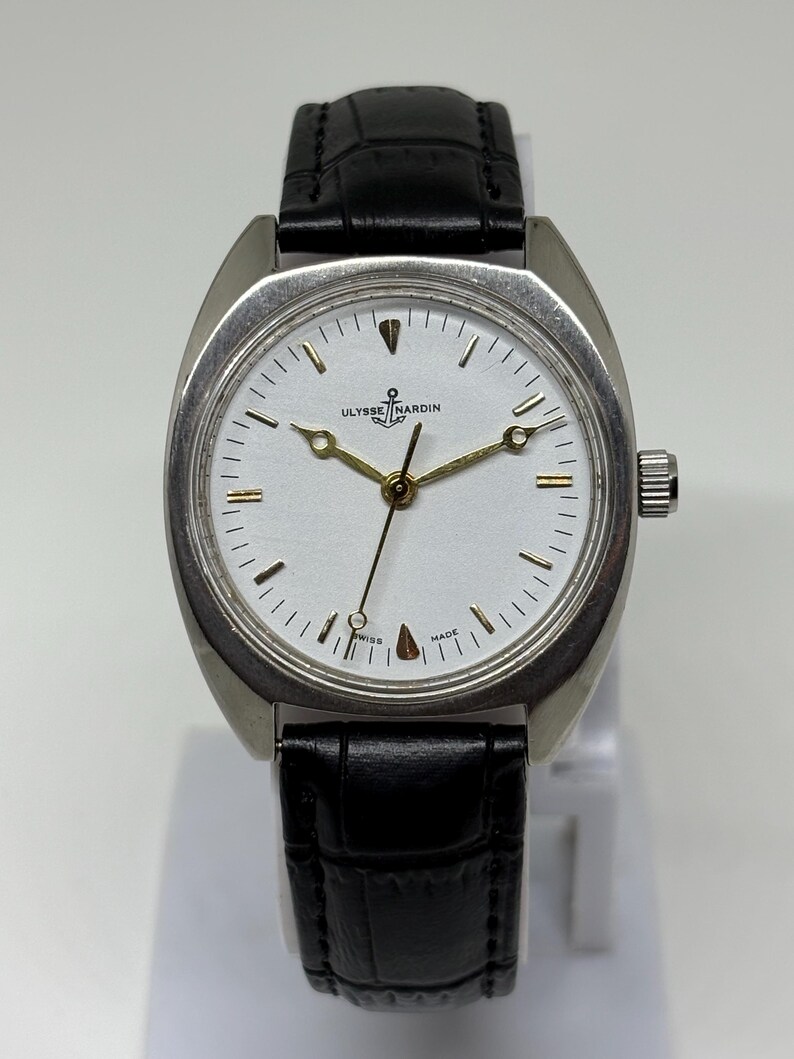 K&ouml;nnte beinhalten: Eine silberfarbene Armbanduhr mit wei&szlig;em Zifferblatt, goldfarbenen Zeigern und einem Anker-Logo &uuml;ber dem Schriftzug Ulysse Nardin. Die Uhr verf&uuml;gt &uuml;ber ein schwarzes Lederarmband mit strukturierter Oberfl&auml;che und eine silberne Krone.