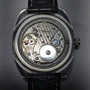 May include: A silver-toned wristwatch with a black leather band. The watch's back is open, revealing the intricate mechanical movement with gears, jewels, and the inscription "SWISS MADE". The watch has a rectangular face and a small crown.