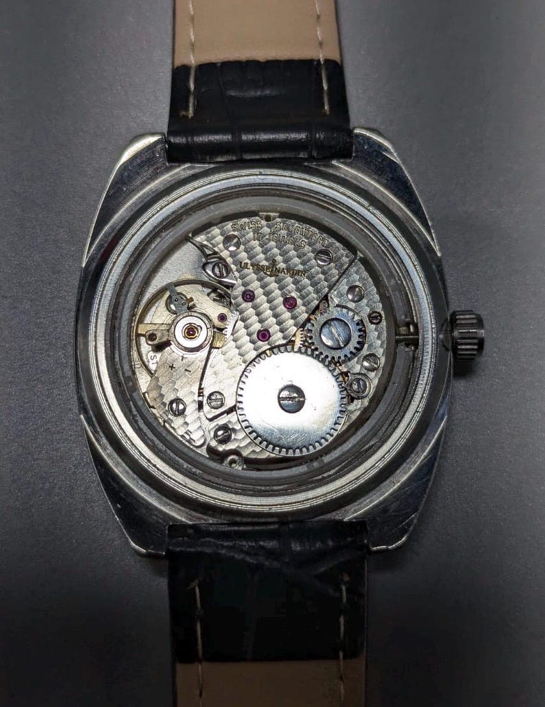 May include: A silver-toned wristwatch with a black leather band. The watch's back is open, revealing the intricate mechanical movement with gears, jewels, and the inscription "SWISS MADE". The watch has a rectangular face and a small crown.