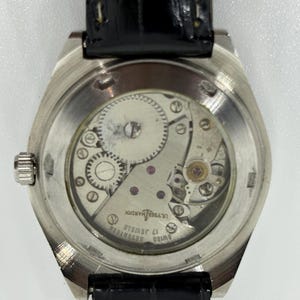 Puede incluir: Un reloj de pulsera plateado con correa de cuero negro y fondo de caja transparente que muestra el movimiento mec&aacute;nico. El movimiento tiene grabadas las palabras Ulysse Nardin, Swiss y Seventeen 17 Jewels. Una peque&ntilde;a corona plateada est&aacute; al lado.