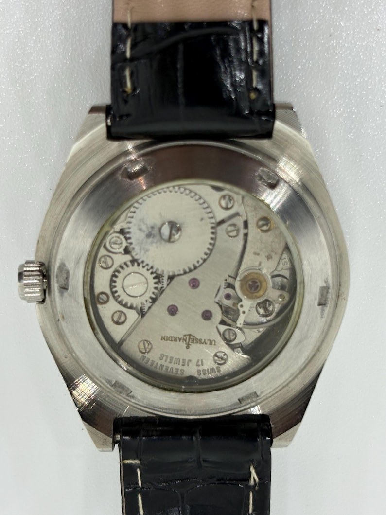 Puede incluir: Un reloj de pulsera plateado con correa de cuero negro y fondo de caja transparente que muestra el movimiento mec&aacute;nico. El movimiento tiene grabadas las palabras Ulysse Nardin, Swiss y Seventeen 17 Jewels. Una peque&ntilde;a corona plateada est&aacute; al lado.