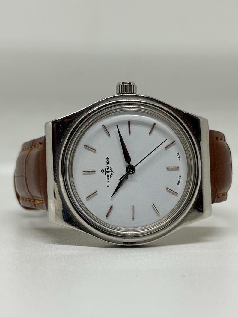 May include: A silver-tone wristwatch with a round white dial, featuring black hands and hour markers. The dial displays the Ulysse Nardin logo with an anchor symbol. It is fitted with a brown leather strap with a textured, embossed pattern.