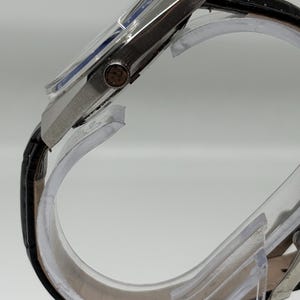 May include: A side view of a silver-toned wristwatch featuring a black leather strap with a textured finish. The watch case is metallic with a prominent winding crown on the side, and the dial is partially visible under a clear, slightly domed crystal.