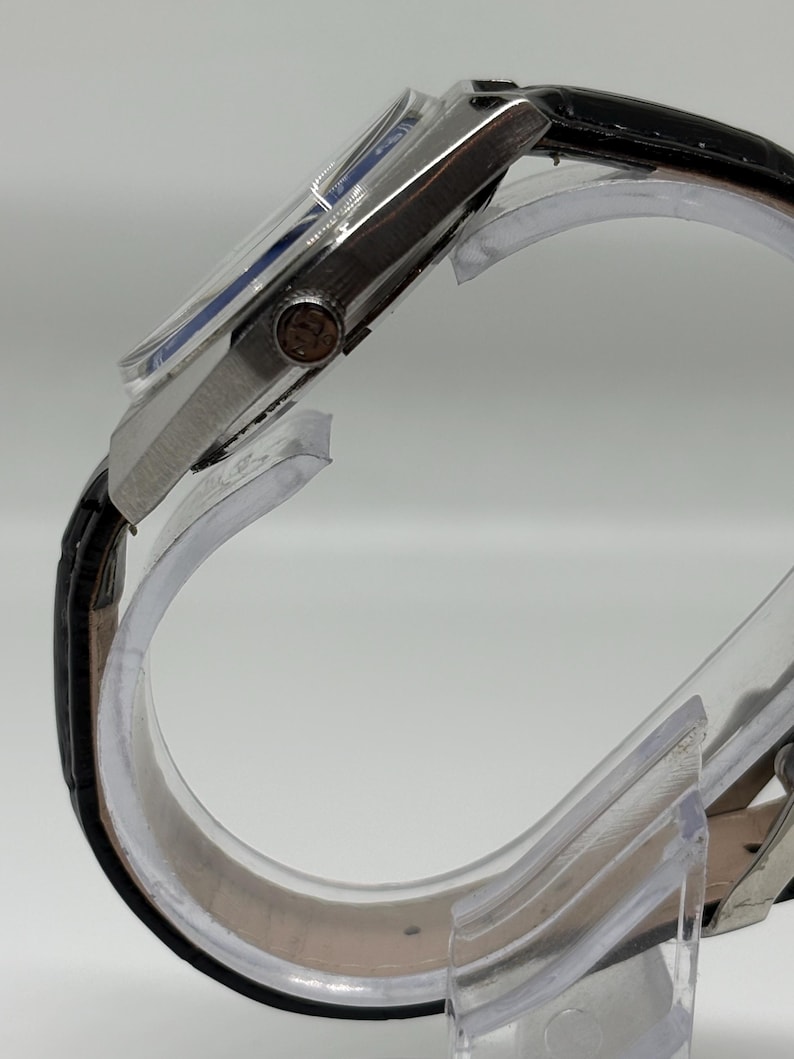 May include: A side view of a silver-toned wristwatch featuring a black leather strap with a textured finish. The watch case is metallic with a prominent winding crown on the side, and the dial is partially visible under a clear, slightly domed crystal.