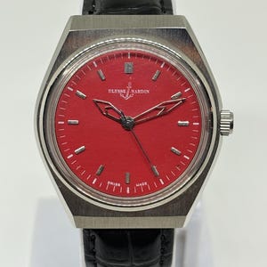 Puede incluir: Un reloj de pulsera Ulysse Nardin con esfera roja vibrante, marcadores de hora plateados y manecillas caladas. El reloj cuenta con una caja de acero inoxidable cepillado y una correa de cuero negro con un patr&oacute;n texturizado de cocodrilo.
